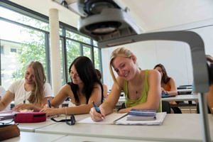 Drei Studentinnen sitzen in der ersten Reihe und schreiben auf Blöcke. Sie lachen und haben Spaß.