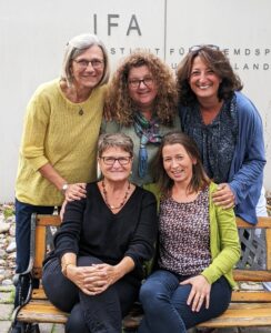 3 stehende Frauen lachen in die Kamera, zwei sitzen auf einer Bank davor und lachen ebenfalls