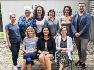 5 lachende Frauen stehend und ein Mann rechts stehend, davor auf einer Bank sitzend drei lächelnde Frauen